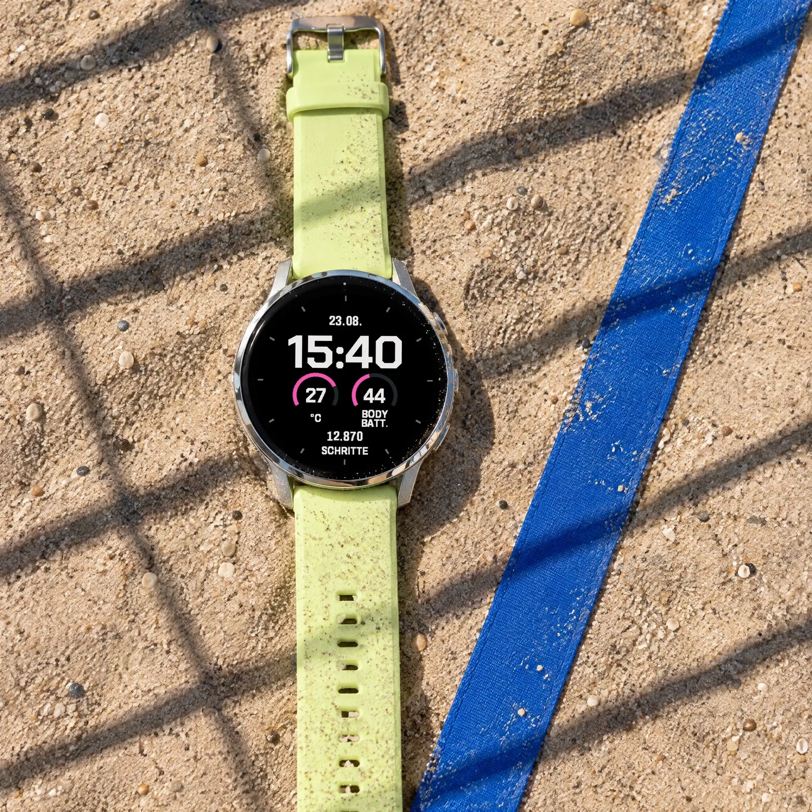 Garmin Venu 4 mit dem Fieldline Garmin Watch Face in Grauweiß / Rosa im Sand eines Beachvolleyballfelds