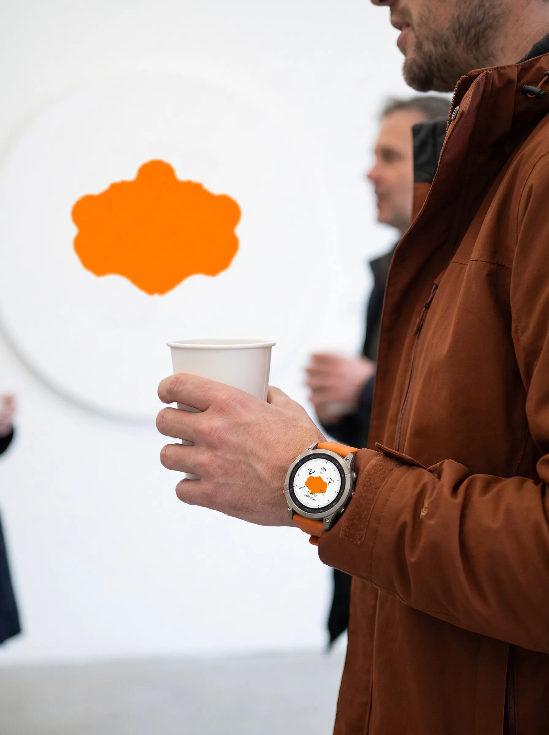 Garmin fēnix 9 Pro mit dem Watch Face Ulm in Orange am Handgelenk, in der Hand ein Kaffeebecher